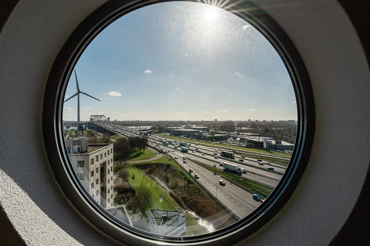 Uitzicht door een rond raam op de snelweg bij Rivium Quadrant met een windmolen, brug en zonlicht aan de horizon.